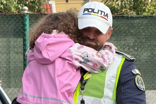 Un bărbat cu șapcă albă pe care scrie POLITIA îmbrăcat în uniformă și vestă reflectorizantă îmbrățișează un copil cu o haină roz cu desene.