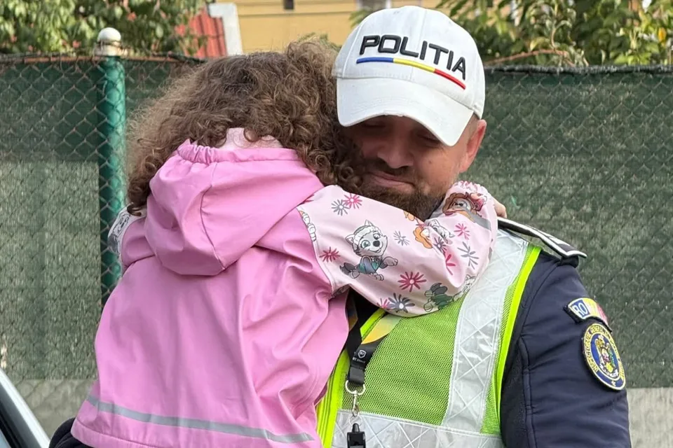Un bărbat cu șapcă albă pe care scrie POLITIA îmbrăcat în uniformă și vestă reflectorizantă îmbrățișează un copil cu o haină roz cu desene.
