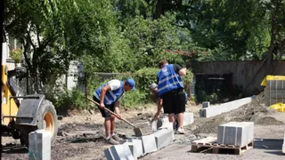 Unii lucrători cu veste albastră și căciulcă portocalie manipulează blocuri de beton pe un teren de șantier cu utilaje și arbori în fundal.