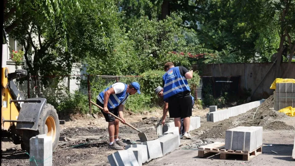 Unii lucrători cu veste albastră și căciulcă portocalie manipulează blocuri de beton pe un teren de șantier cu utilaje și arbori în fundal.