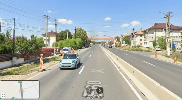 O mașină de culoare deschisă circulă pe un drum cu două benzi, lângă un parapet de beton și un indicator de circulație.