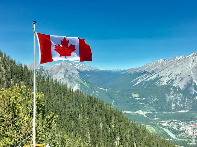 O steag canadian flutură pe un stâlp alb sub un cer senin, deasupra unui peisaj montan cu păduri de conifere și un oraș în vale.