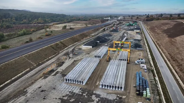 O vedere aeriană prezintă o zonă de șantier cu elemente de beton gri așezate pe sol, lângă o porțiune de drum asfaltat și vegetație.