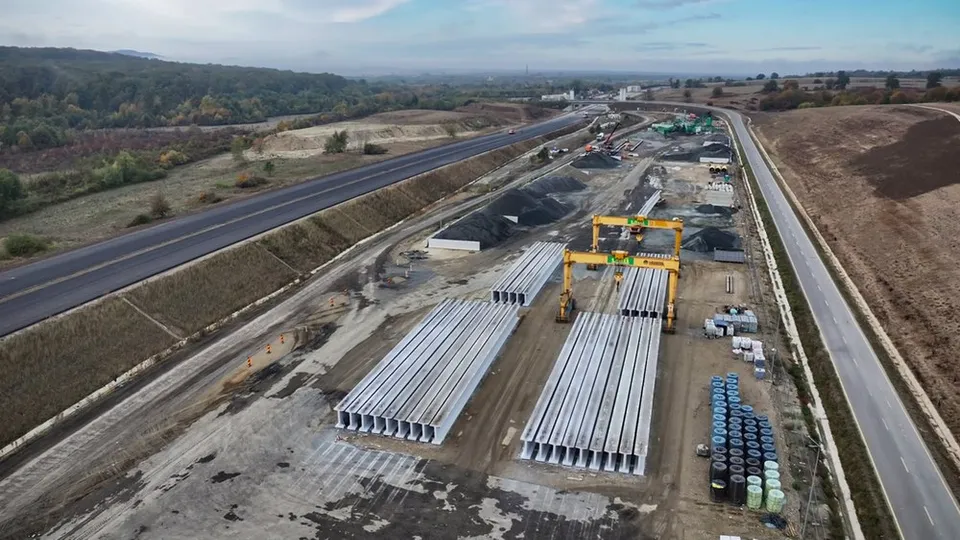 O vedere aeriană prezintă o zonă de șantier cu elemente de beton gri așezate pe sol, lângă o porțiune de drum asfaltat și vegetație.