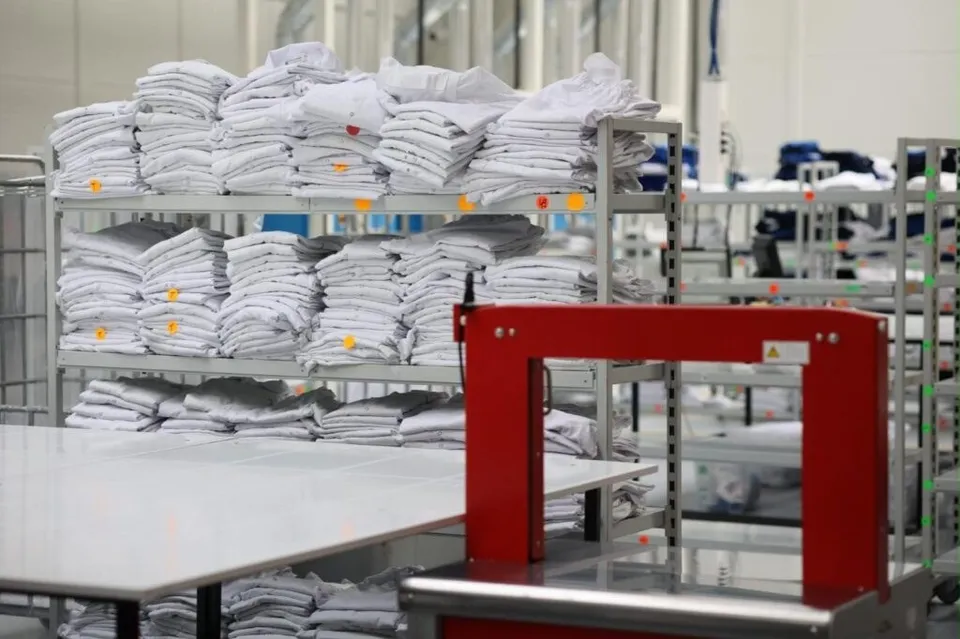Pachete cu haine albe sunt stivuite pe rafturi metalice într-un spațiu industrial lângă o masă albă și un echipament roșu.