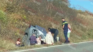 Un autoturism gri răsturnat se află pe un taluz de iarbă lângă un drum, în timp ce mai mulți oameni stau lângă vehicul.