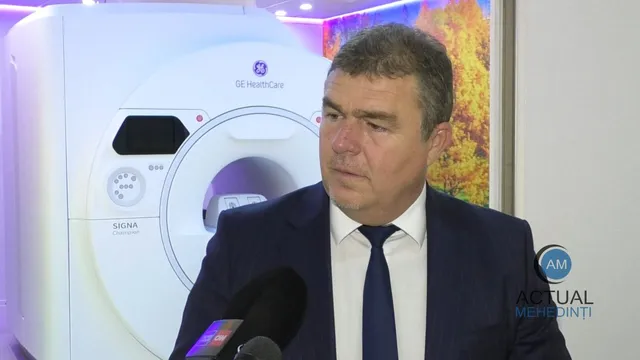 Un bărbat într-un costum închis și cămașă albă cu cravată stă în fața unui aparat medical alb de tip RMN.