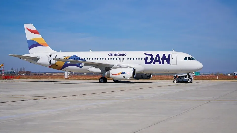 Un avion alb cu inscripția DAN și elemente colorate pe fuselaj și coadă stă pe o pistă de beton sub un cer senin.