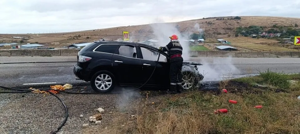 Un pompier cu cască portocalie și echipament de protecție stă lângă un autoturism negru de care se ridică fum pe marginea unui drum asfaltat.