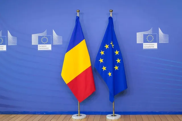 Două steaguri, cel al României și cel al Uniunii Europene, sunt expuse pe suporturi metalice în fața unui fundal albastru cu embleme albe.