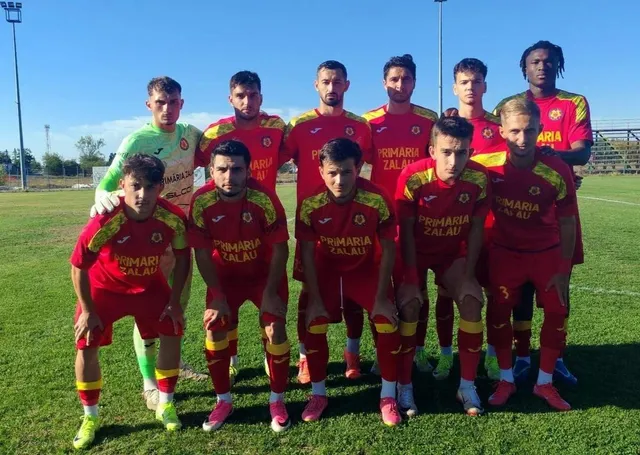 O echipă de fotbaliști în echipamente roșii și unul în echipament verde stau grupați pe un teren de fotbal sub cerul senin.