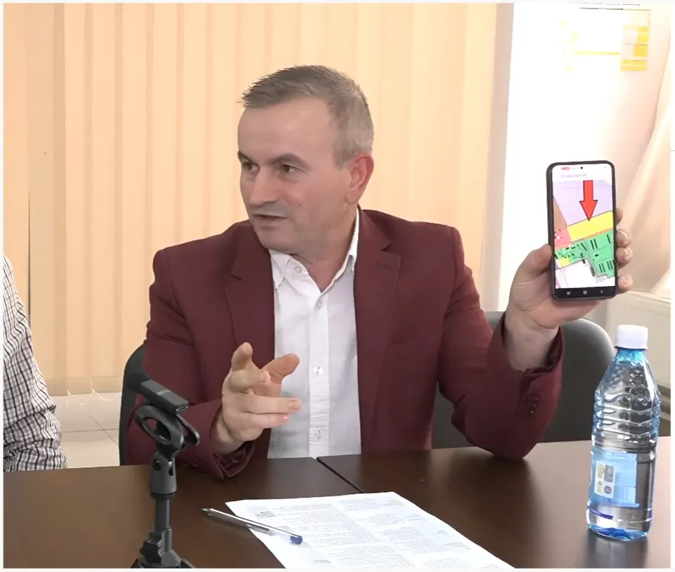 Un bărbat într-un sacou roșu stă la o masă și ține un telefon mobil care afișează o hartă cu o săgeată roșie.