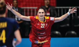 O femeie cu mâinile întinse poartă un tricou roșu cu numărul 27 și este pe un teren de sport.