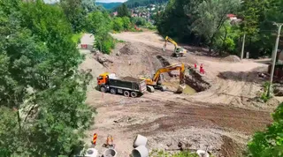 Două excavatoare galbene și un camion negru cu cabină portocalie lucrează pe un teren de pământ deschis, înconjurat de vegetație abundentă.