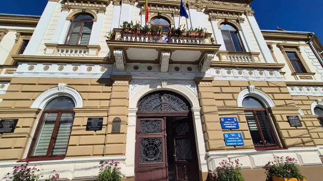O clădire cu fațadă galbenă prezintă ferestre cu rame maro, un balcon cu flori și un pridvor central cu uși de lemn închisă parțial.