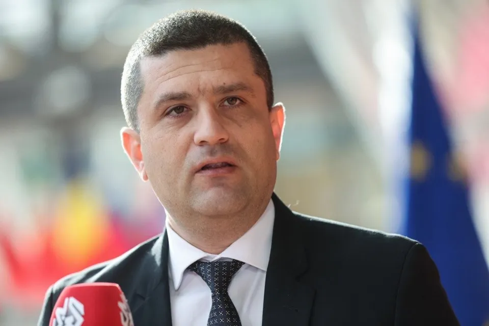 Un bărbat cu părul scurt, purtând cămașă albă, cravată neagră și sacou întunecat, este surprins într-un prim-plan cu un microfon în partea stângă.
