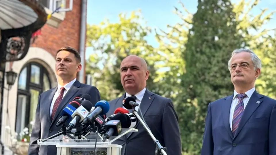 Trei bărbați în costume și cravate stau în spatele unui podium cu mai multe microfoane în fața unor copaci verzi și a unei clădiri de cărămidă.