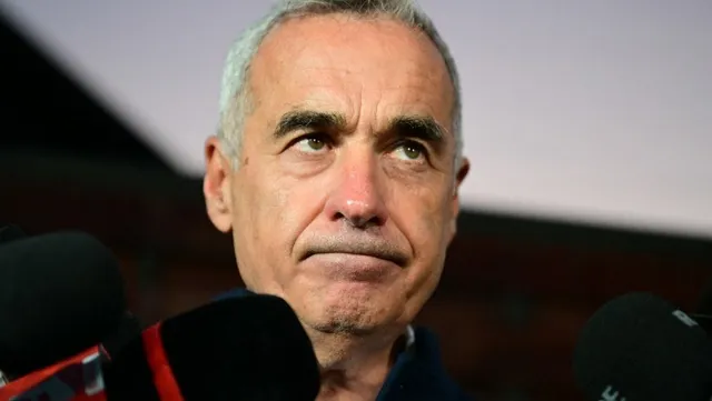 Un bărbat cu părul scurt și cărunt privește în sus, fiind înconjurat de microfoane negre în prim-plan.