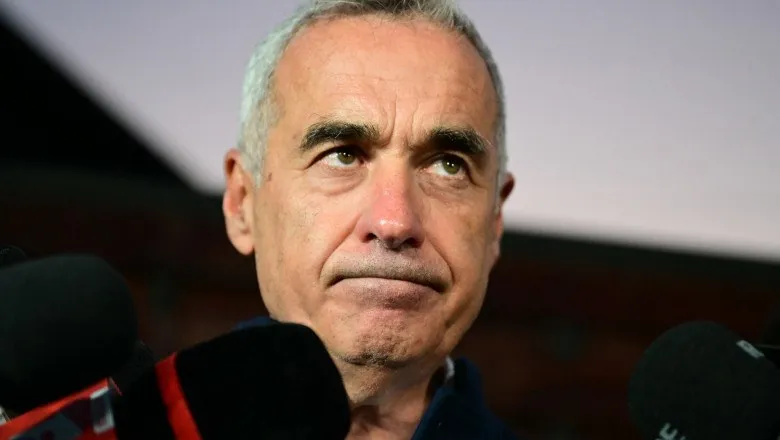 Un bărbat cu părul scurt și cărunt privește în sus, fiind înconjurat de microfoane negre în prim-plan.