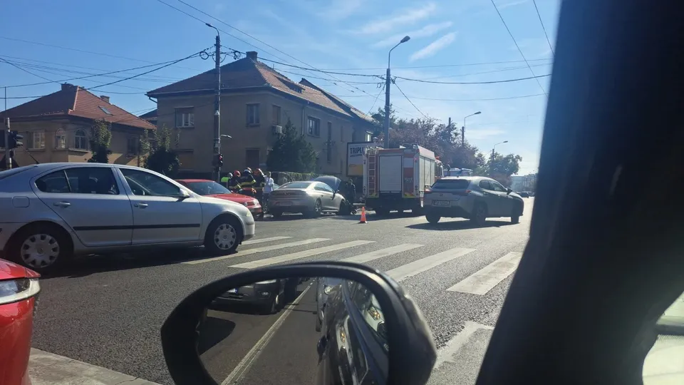 O mașină gri și un camion alb sunt parcate pe un drum asfaltat lângă clădiri sub un cer senin.