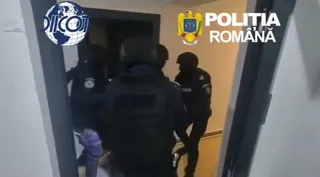 Patru persoane purtând uniforme închise și echipament tactice se află într-un hol lângă o ușă deschisă.