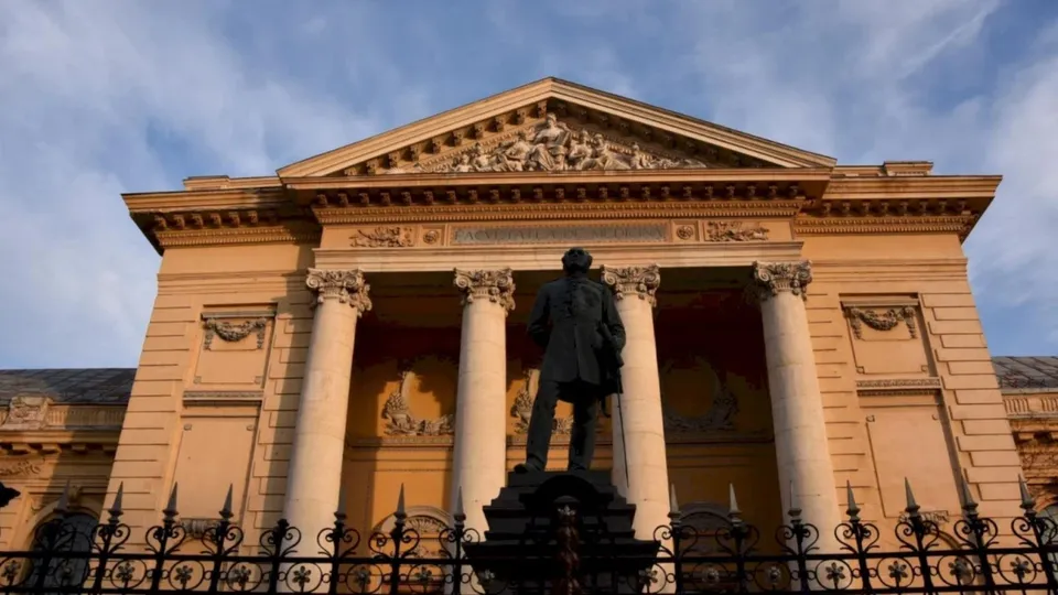 O statuie neagră de spate în fața unei clădiri clasice cu coloane și un fronton sculptat sub un cer înnorat.