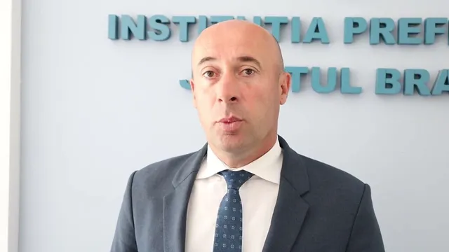 Un bărbat în costum de afaceri gri cu cravată albastră cu puncte albe, în picioare în fața unei pereți albe cu litere albastre.