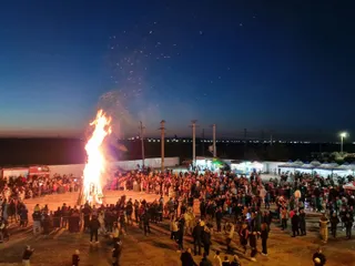 O mulțime mare de oameni este adunată în jurul unui foc mare, într-un spațiu deschis pe parcursul nopții, sub un cer încețurat de stele.