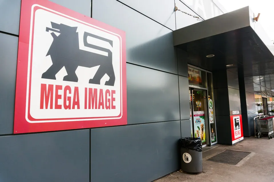 Un panou roșu cu un logo alb și textul Mega Image este montat pe o fațadă de culoare gri lângă o intrare.