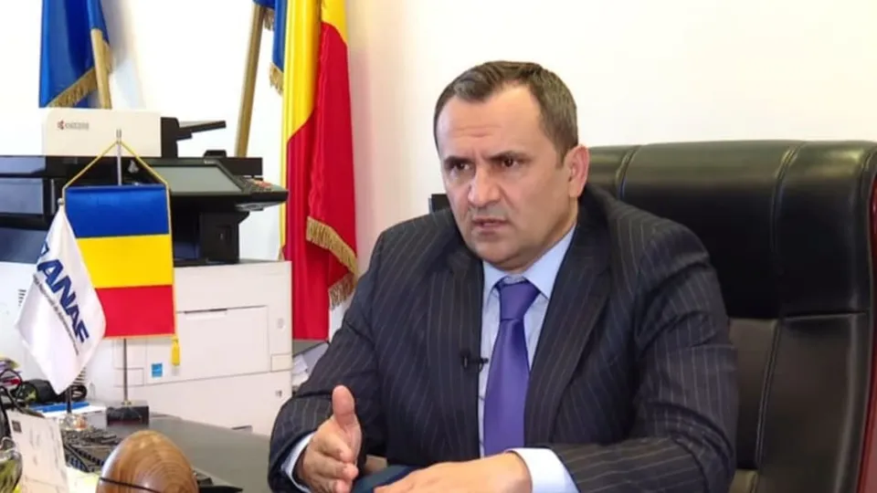 Un bărbat cu sacou în dungi și cravată violet stă la un birou lângă un steag și un aparat de imprimantă.