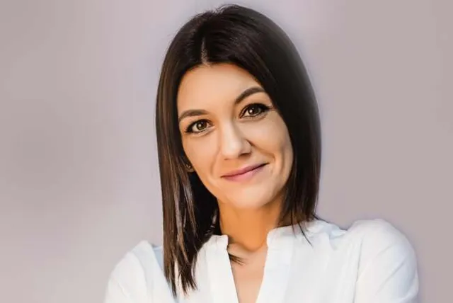 O femeie cu părul scurt și negru poartă o cămașă albă pe un fundal gri deschis.