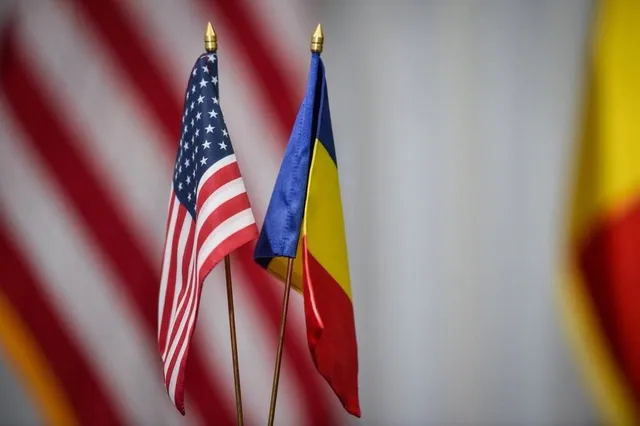 Două steaguri mici, unul american și unul român, sunt montate pe suporturi subțiri de metal pe un fundal neclar.