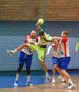 Un jucător în echipament verde sare spre o minge galbenă, fiind contactat de doi jucători în echipamente alb-roșii și albastre într-o sală de sport.