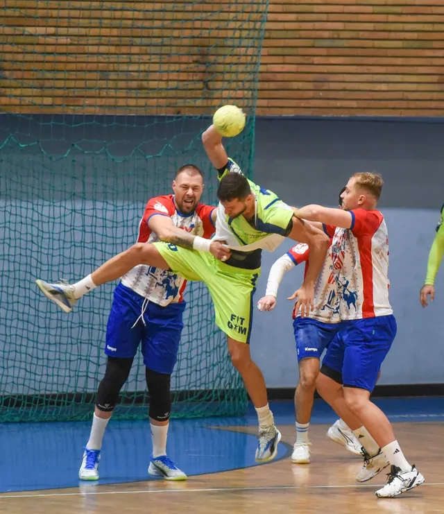 Câțiva jucători de handbal în echipamente colorate se află pe un teren de sport în fața unei ținte cu plasă.