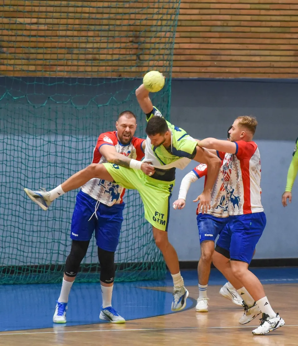 Câțiva jucători de handbal în echipamente colorate se află pe un teren de sport în fața unei ținte cu plasă.