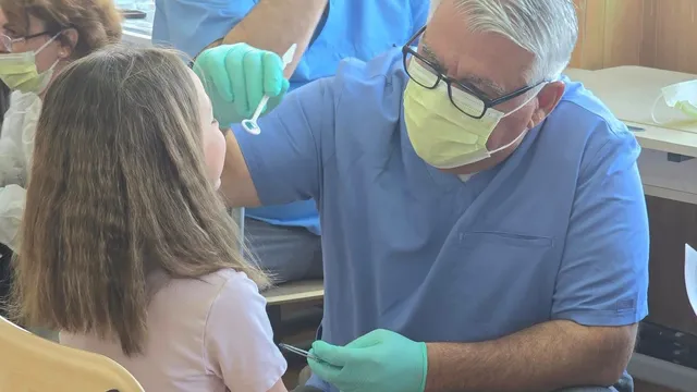 Un stomatolog cu mască și mănuși verzi examinează dinții unei fete cu părul lung într-un cabinet medical.