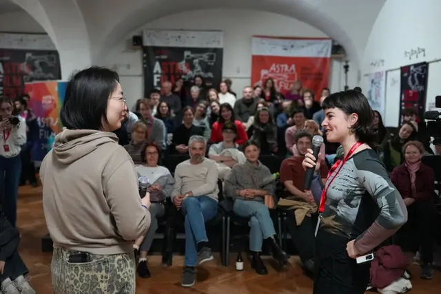 O femeie cu păr scurt și tricou gri ține un microfon, în timp ce o altă femeie cu hanorac bej o privește într-o sală cu public.