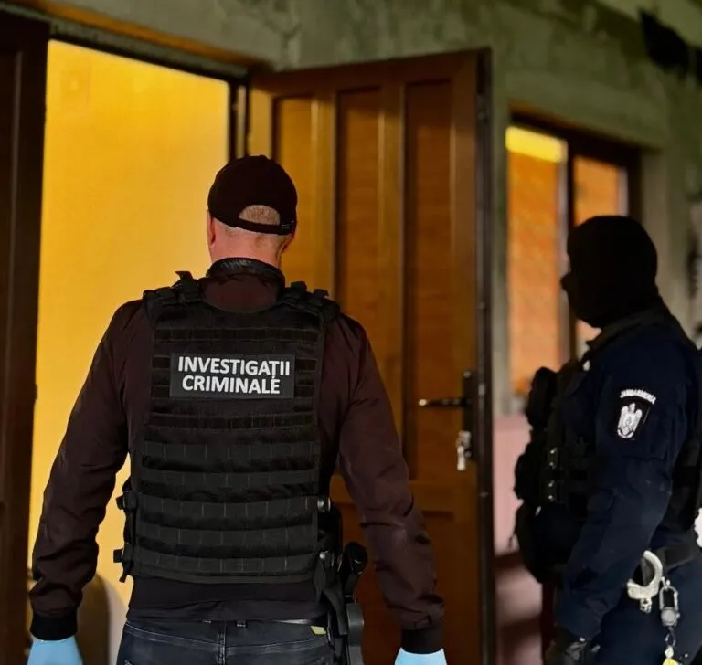 Un bărbat cu vestă tactică pe care scrie INVESTIGATII CRIMINALE stă cu spatele în fața unei uși deschise lângă un alt agent în echipament tactic.