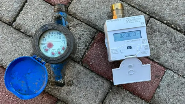 Un contor de apă alb cu un ecran digital și un contor de apă albastru cu un cadran analogic sunt așezate pe pavaj.