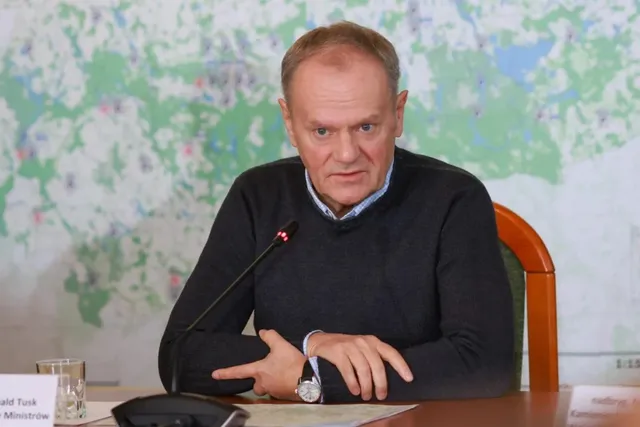 Un bărbat cu părul scurt și cămașă sub un pulover închis la culoare stă la o masă lângă un microfon și un pahar de sticlă.