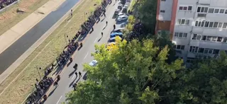 O mulțime de oameni stă la rând pe o stradă lângă un canal, lângă un bloc de locuințe și copaci verzi.