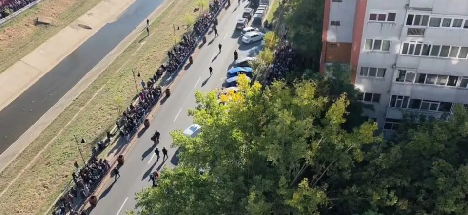O mulțime de oameni stă la rând pe o stradă lângă un canal, lângă un bloc de locuințe și copaci verzi.
