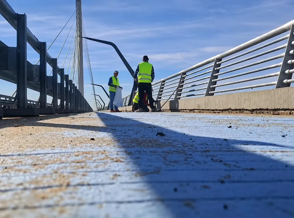 Un bărbat cu vestă reflectorizantă verde și un alt bărbat cu vestă reflectorizantă portocalie lucrează pe o suprafață de culoare deschisă lângă un parapet metalic.