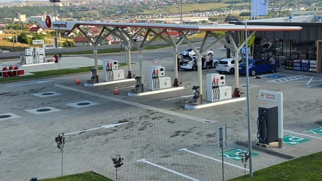 O benzinărie cu pompe de alimentare, un acoperiș susținut de piloni albi și câteva mașini parcate pe un teren pavat cu pietriș gri.