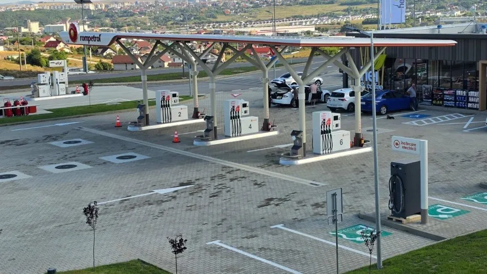 O benzinărie cu pompe de alimentare, un acoperiș susținut de piloni albi și câteva mașini parcate pe un teren pavat cu pietriș gri.