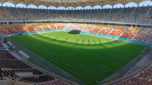 O vedere de sus asupra unui stadion de fotbal cu tribune colorate și un teren de gazon verde sub un acoperiș de tip copertină.
