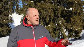 Un bărbat cu părul scurt și pielea deschisă poartă o jachetă gri și roșie, stând în fața unor copaci coniferi acoperiți de zăpadă.