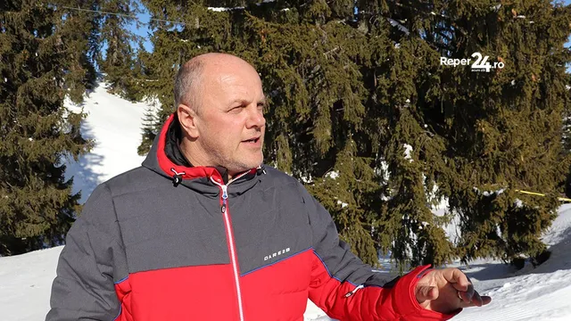 Un bărbat cu părul scurt și pielea deschisă poartă o jachetă gri și roșie, stând în fața unor copaci coniferi acoperiți de zăpadă.