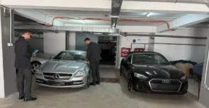 Două mașini sunt parcate într-un garaj subteran, în timp ce doi bărbați stau lângă un autoturism argintiu.