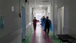 Persoane în halate medicale merg pe un coridor de spital cu pereți albi și podea roz și verde.
